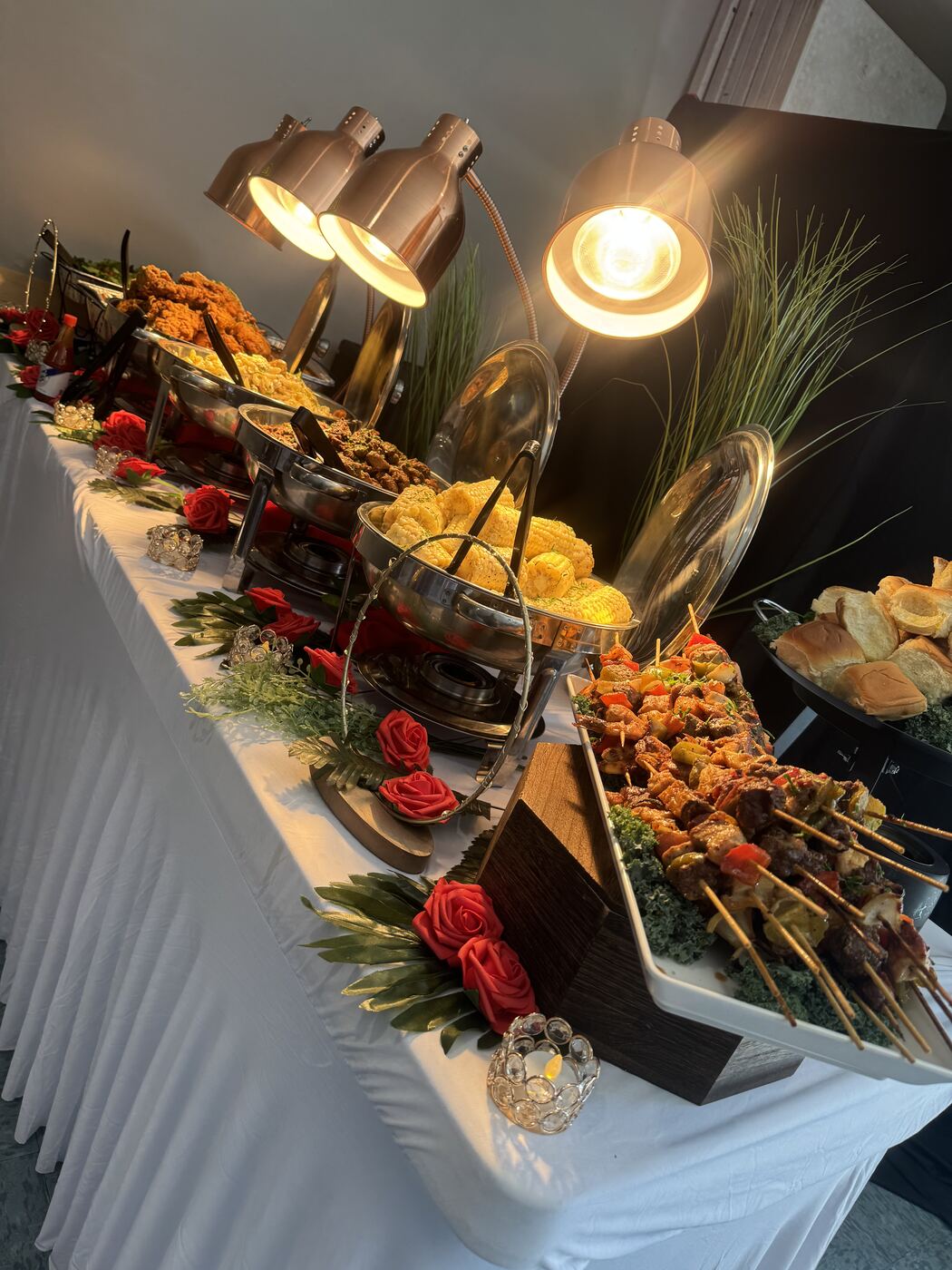 Catering buffet with grilled skewers and fried chicken under heat lamps, styled with roses