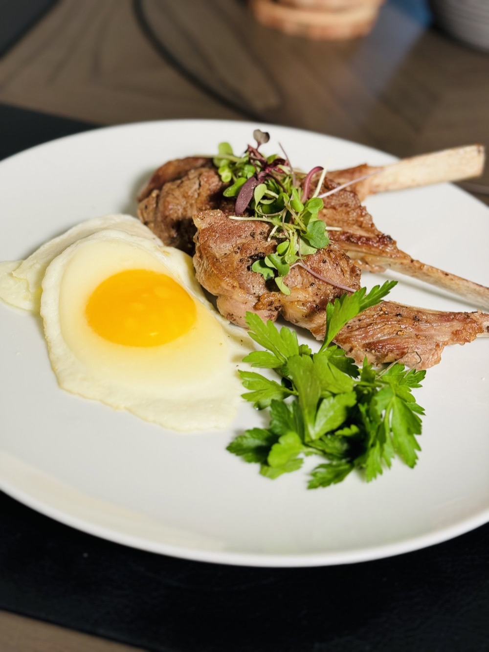 Plated private chef course of frenched lamb chops with a sunny-side egg and microgreens